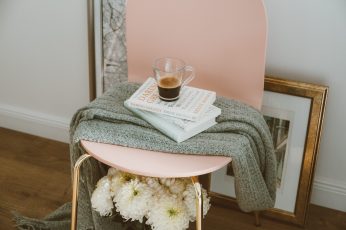 Clear glass mug on three stacked books inside room, pink chair with gray scarf over books and clear glass mug