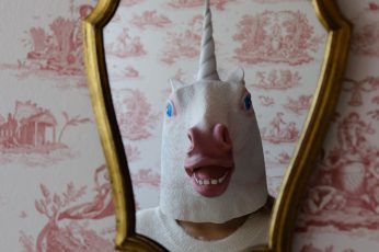 Person with white unicorn mask stands in front of wall mirror wallpaper